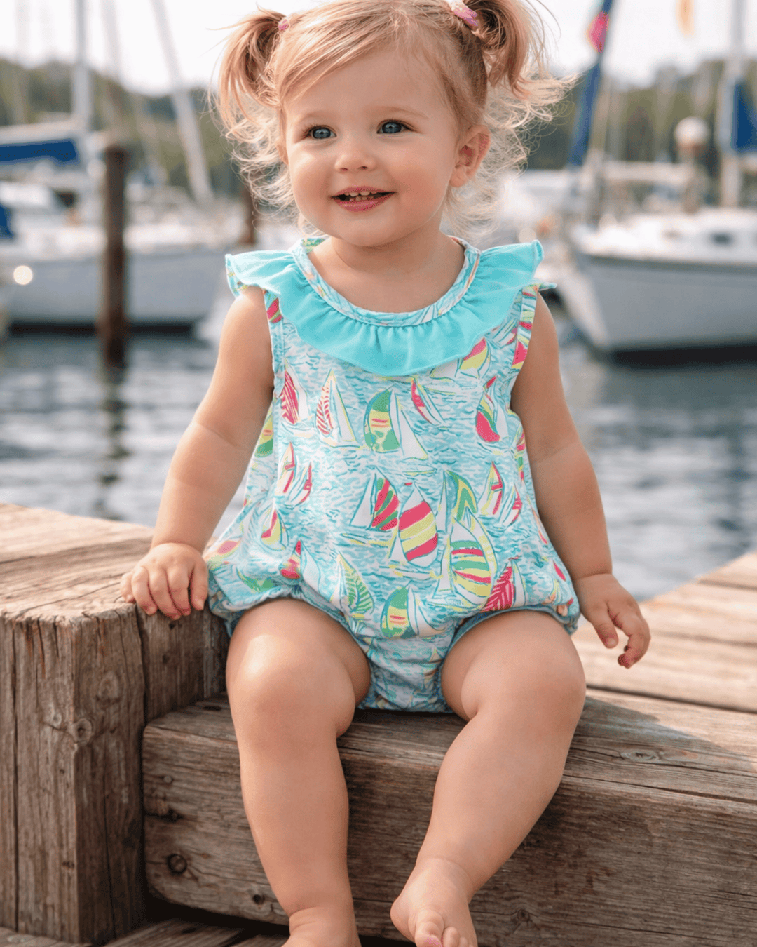 Child wearing a colorful swimsuit with sailboat pattern sitting on a wooden dock by water.