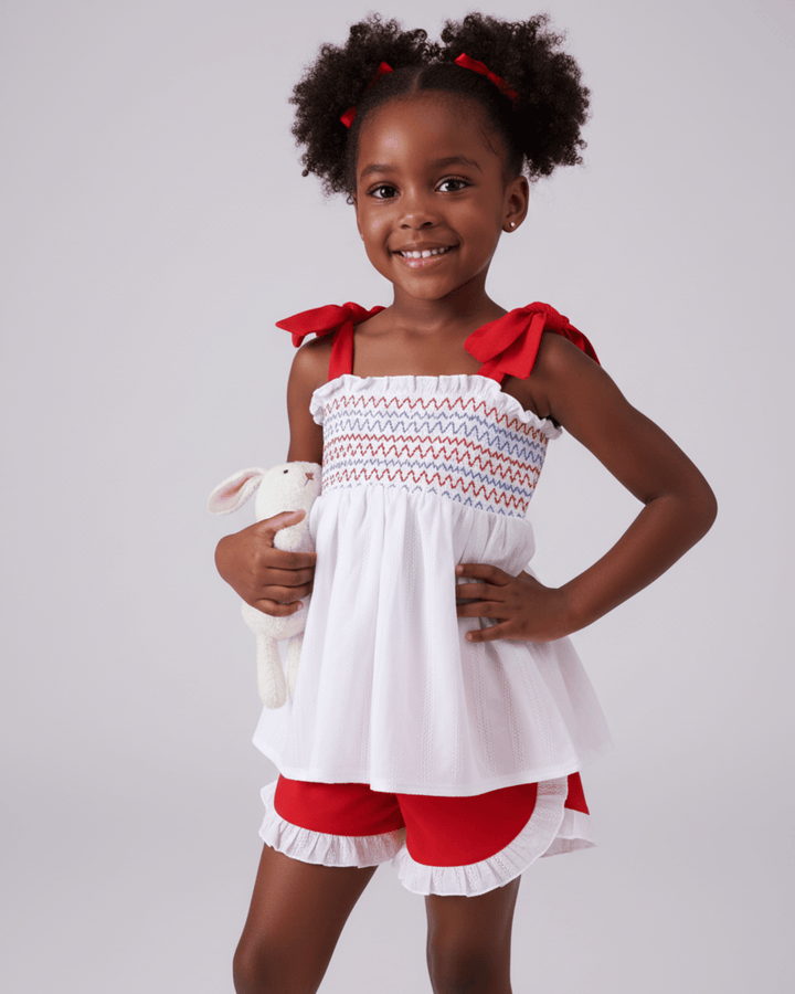 Young girl wearing a white dress with red accents and red shorts, holding a stuffed animal on a gray background.