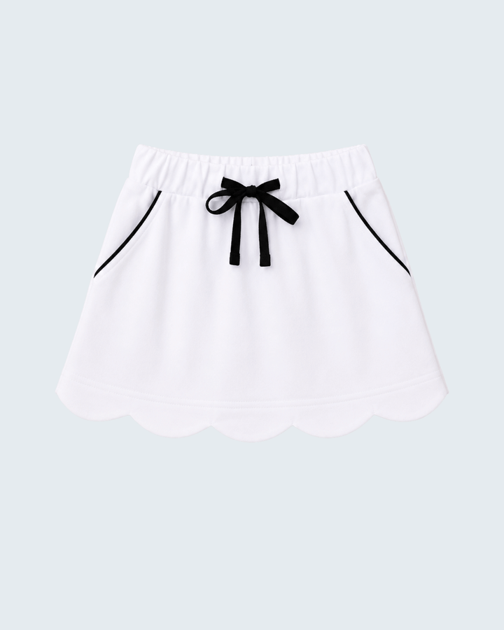 A white skort with a scalloped hem, an elastic waistband, a black ribbon bow at the center, and thin black piping details on the sides, shown against a light gray background.