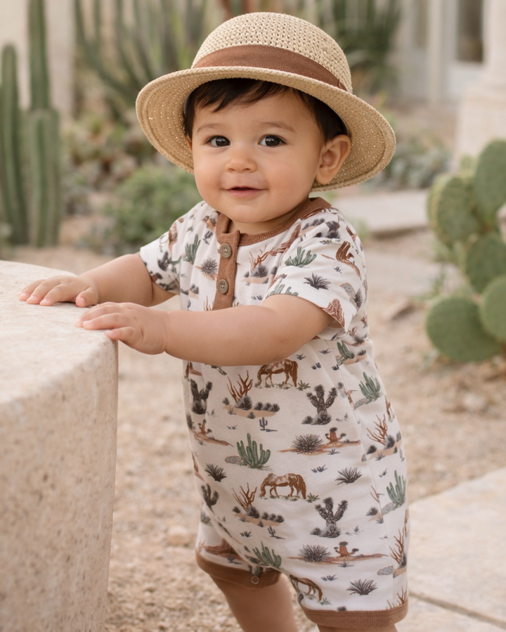 Baby in a desert setting wearing a patterned romper and straw hat.