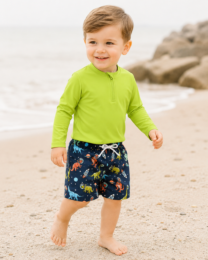 Child wearing a green long-sleeve shirt and dinosaur shorts on a beach.