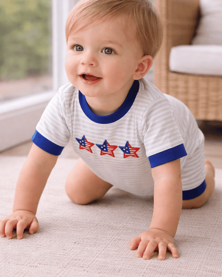 Baby crawling on a carpeted floor wearing a white onesie with blue ringer sleeves and red, white, and blue star designs.