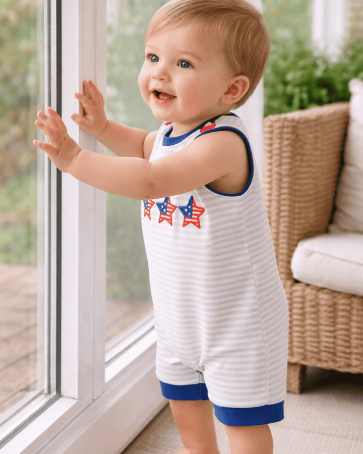 Child wearing a white sleeveless romper with red and blue accents, standing by a window.