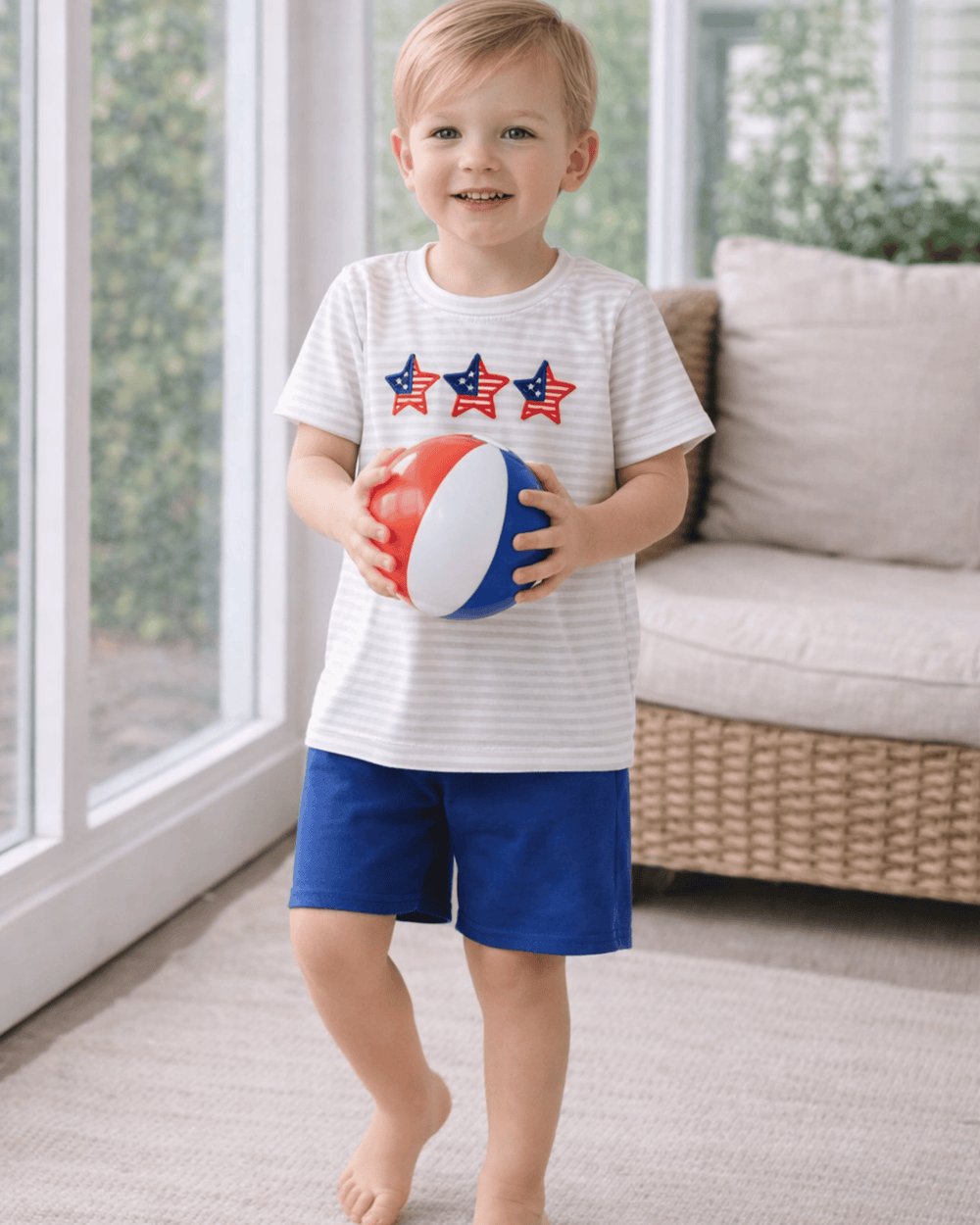 Child holding a beach ball indoors, wearing a striped shirt and blue shorts.