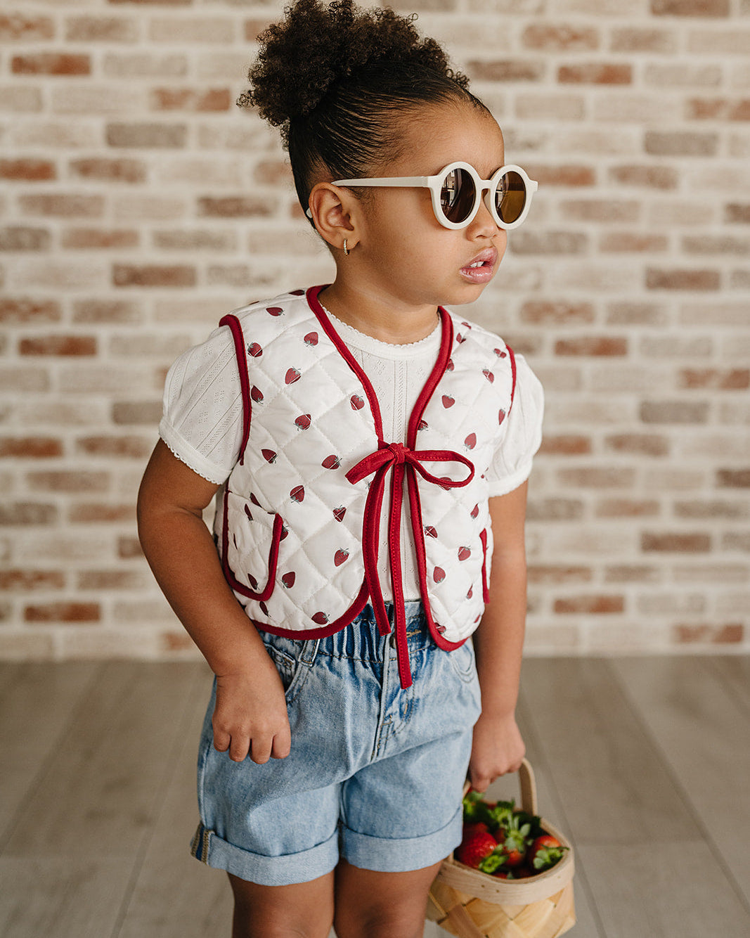 Child wearing a white blouse with red accents and denim shorts, holding a basket against a brick wall.