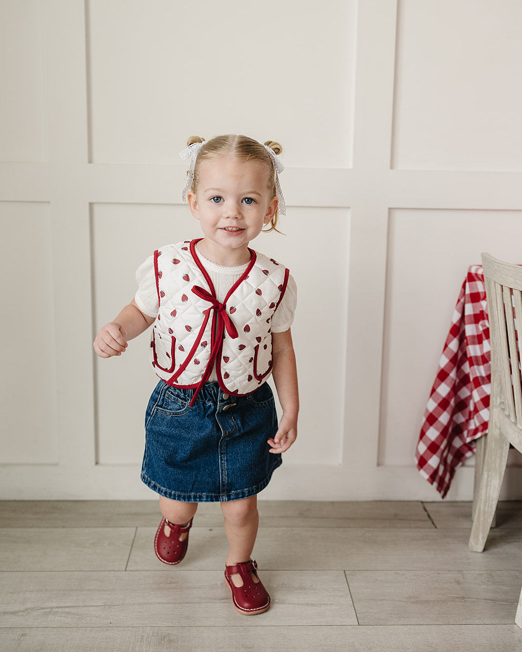 Young girl in a red and white outfit standing in a room with a white wall and wooden floor.