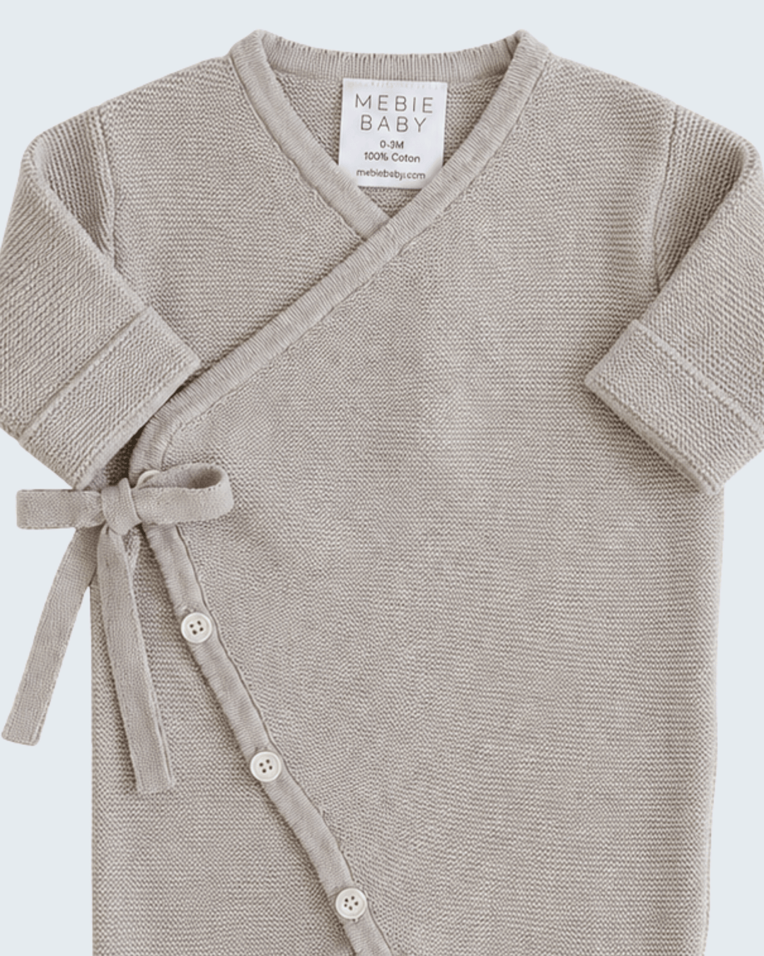 A close-up of a baby wrap top in a light taupe-beige knit fabric, featuring a V-neck, short sleeves, and a side tie with snap buttons. The brand label reads &