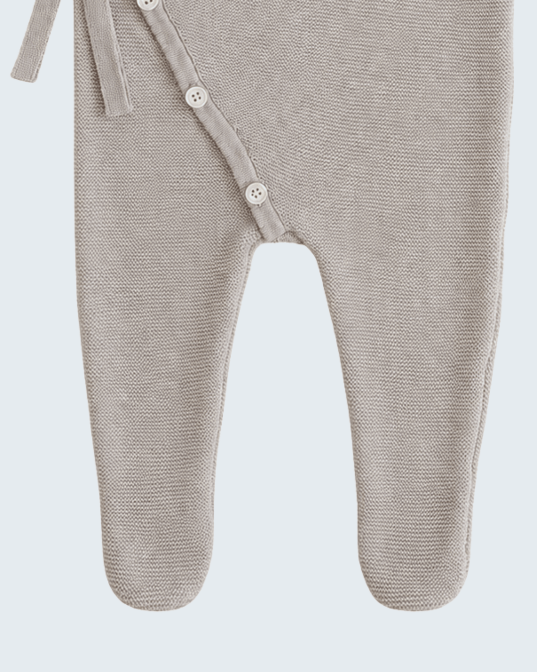 Close-up of a baby footed sleeper in a light taupe-beige knit fabric, showing the side snap closure with white buttons and a tie strap against a light gray background.