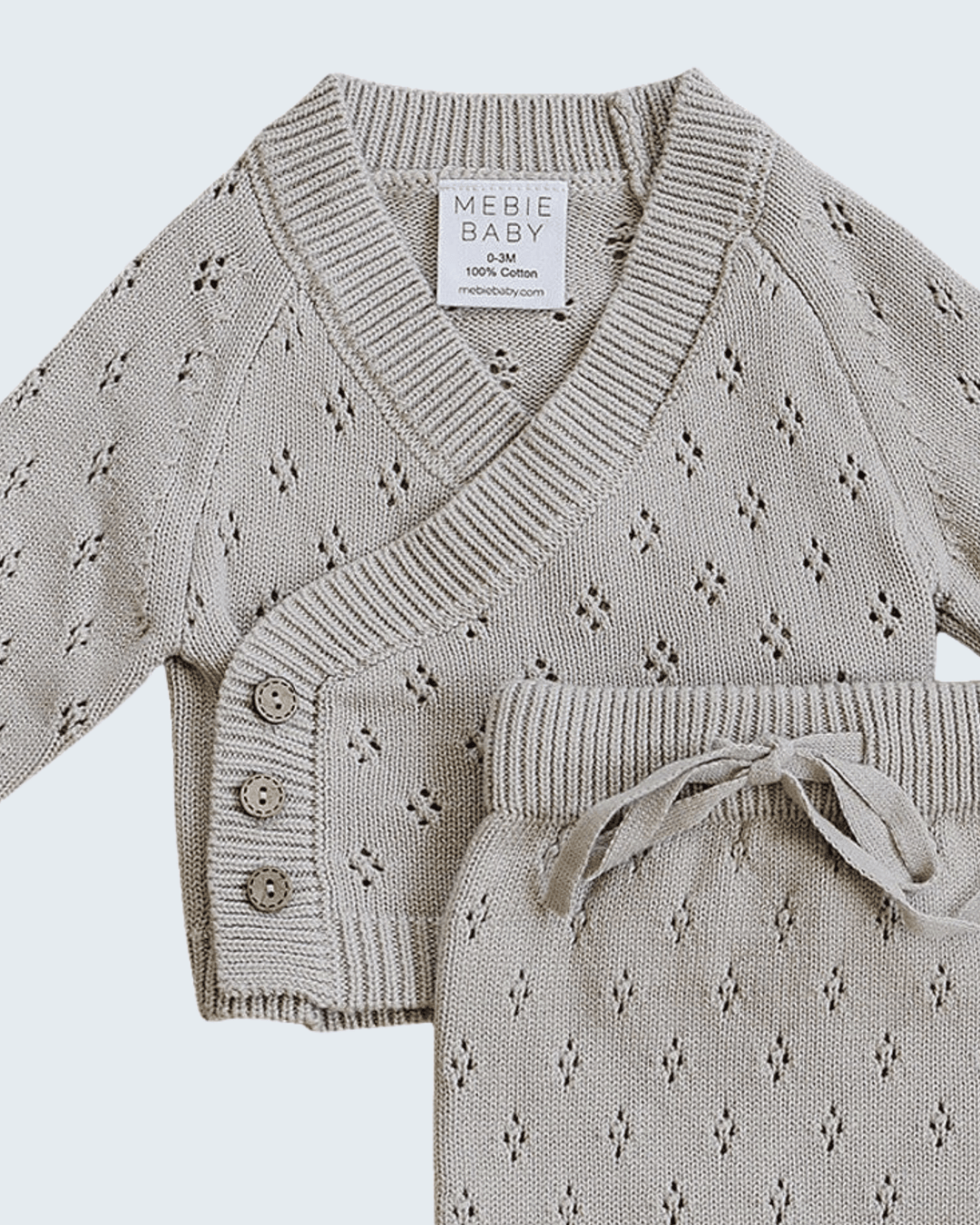 A baby knit two-piece set in a light oatmeal beige color, featuring a V-neck wrap cardigan with button closures and matching knit pants with a drawstring waist, shown on a light gray background.