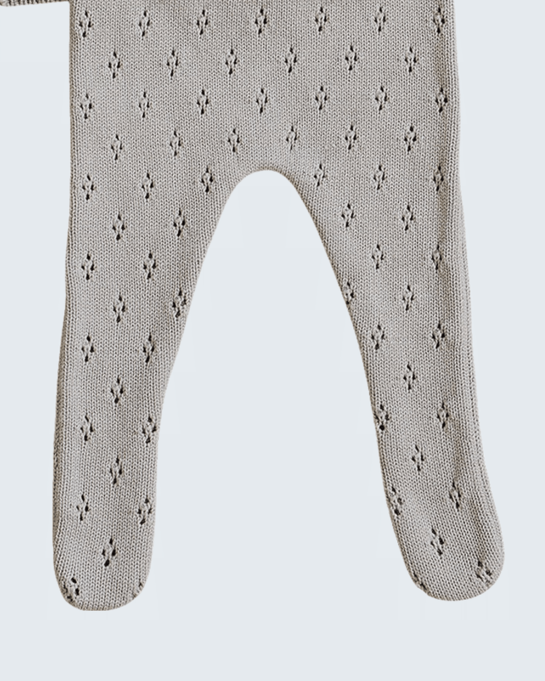 A close-up of a knit baby bootie or footed sock in a light taupe-beige color with a small repeating dark geometric pattern, shown against a light gray background.