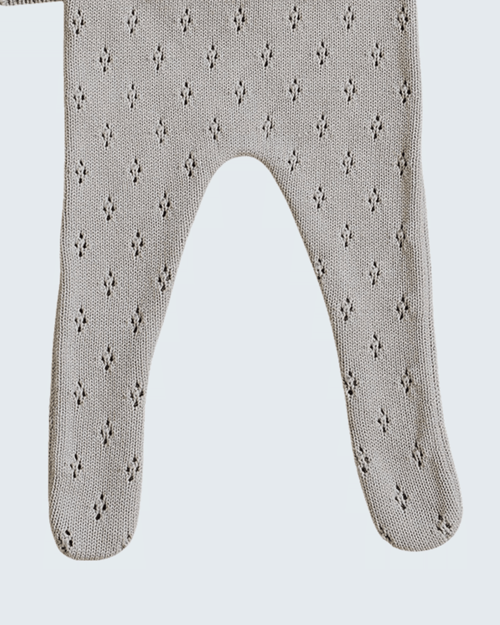 A close-up of a knit baby bootie or footed sock in a light taupe-beige color with a small repeating dark geometric pattern, shown against a light gray background.