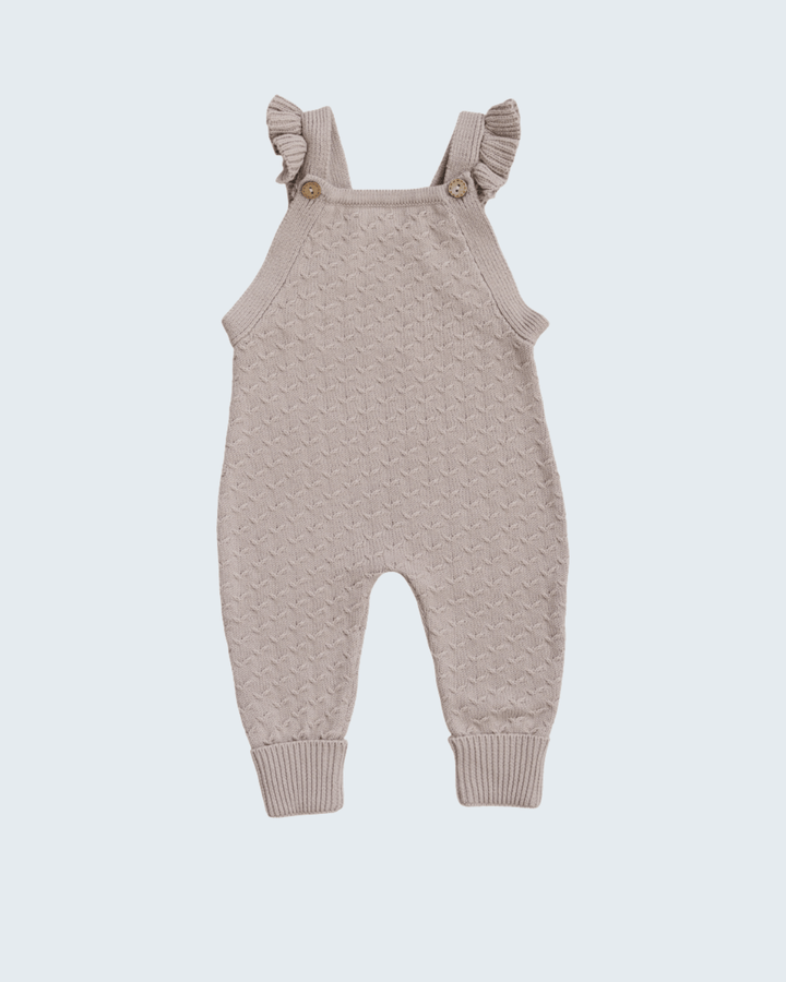 A light beige knit baby jumpsuit with ruffled shoulder straps and small wooden buttons, shown against a light gray background.