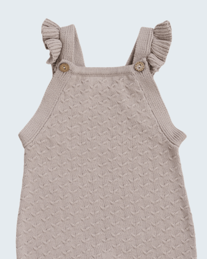 A close-up of a baby knit romper in a light taupe-beige color, featuring textured knit fabric, ruffled shoulder straps, and two wooden buttons.