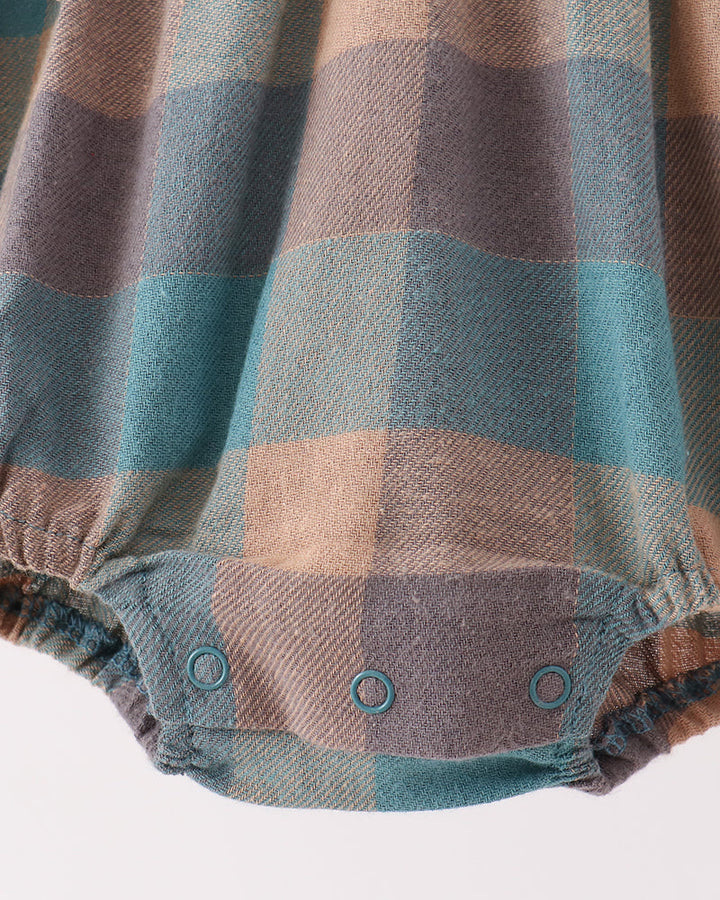 Close-up of a plaid baby bodysuit or romper fabric with three snap closures at the crotch. The pattern features blocks of muted teal blue, dusty purple, and warm tan/beige.