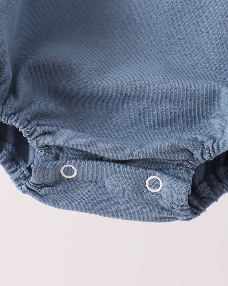 Close-up of a baby romper or bodysuit in dusty blue fabric, showing the elasticized leg openings and two white snap closures on a light off-white background.