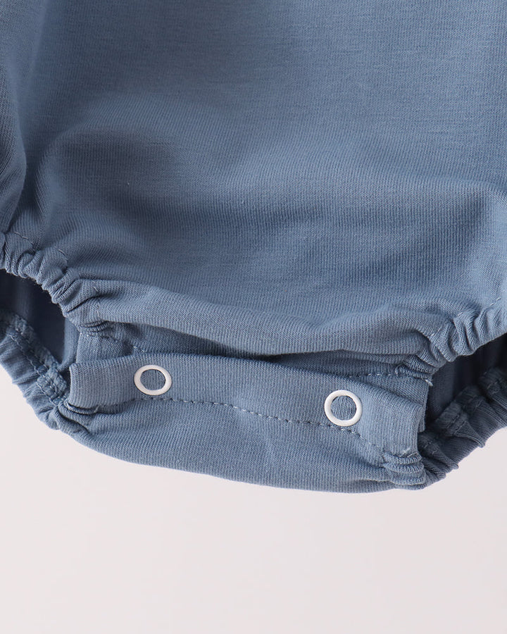 Close-up of a baby romper or bodysuit in dusty blue fabric, showing the elasticized leg openings and two white snap closures on a light off-white background.