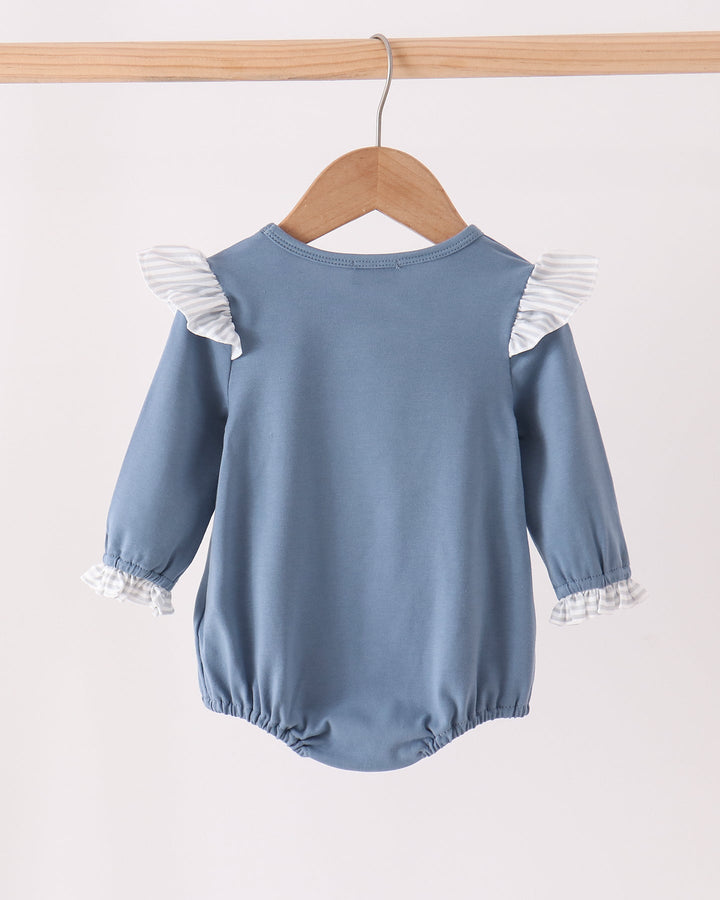 A dusty blue long-sleeve baby romper with white ruffle details on the shoulders and cuffs, hanging on a wooden hanger against a plain off-white background.