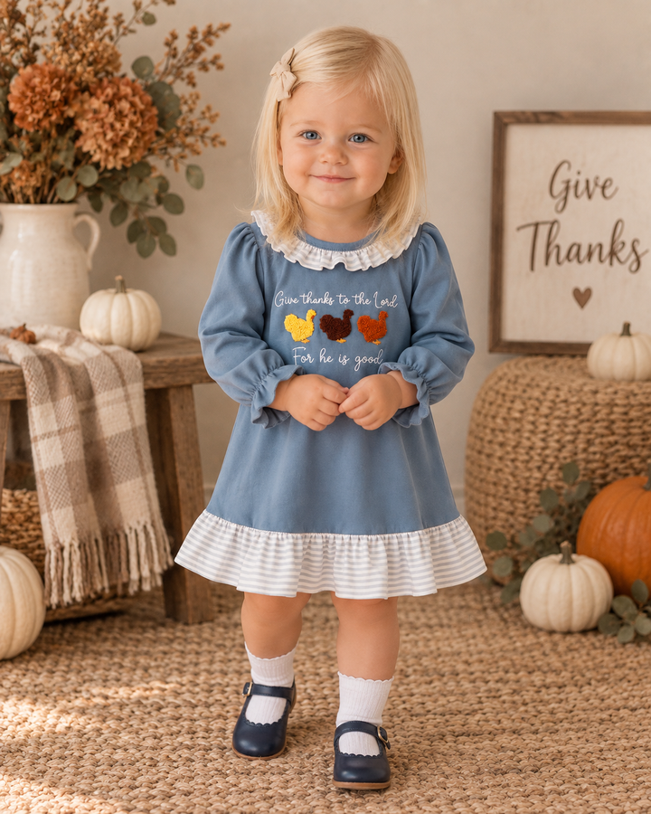 A young toddler girl stands in a cozy autumn-themed room wearing a light blue long-sleeve dress with a white ruffled collar and hem, featuring a turkey graphic on the chest. She is styled with white socks and navy blue shoes, surrounded by pumpkins, a plaid throw blanket, and a rustic wooden sign that reads &