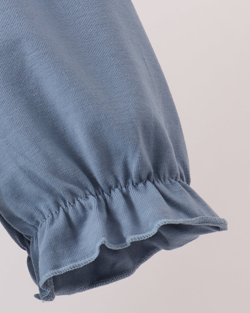 Close-up of a dusty blue fabric sleeve with a gathered ruffled cuff, showing the texture and stitching against a light beige background.
