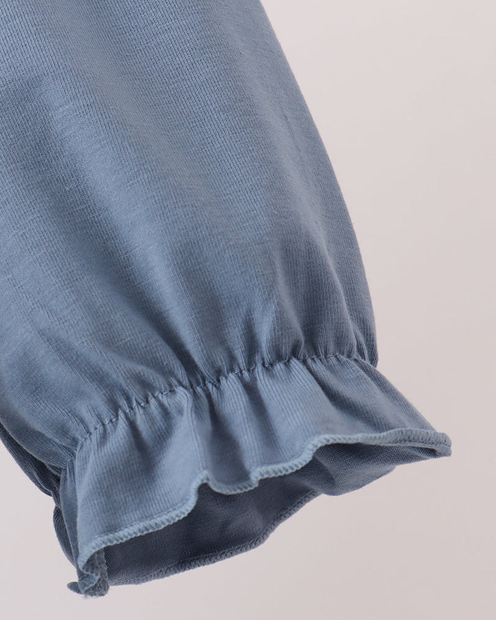 Close-up of a dusty blue fabric sleeve with a gathered ruffled cuff, showing the texture and stitching against a light beige background.