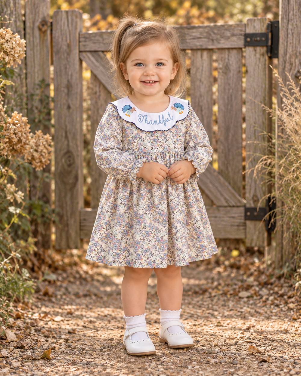 A toddler girl stands outdoors in front of a weathered wooden gate and fence, wearing a long-sleeve floral dress with a white collar that says 'Thank you!' in blue script, white socks, and white shoes.