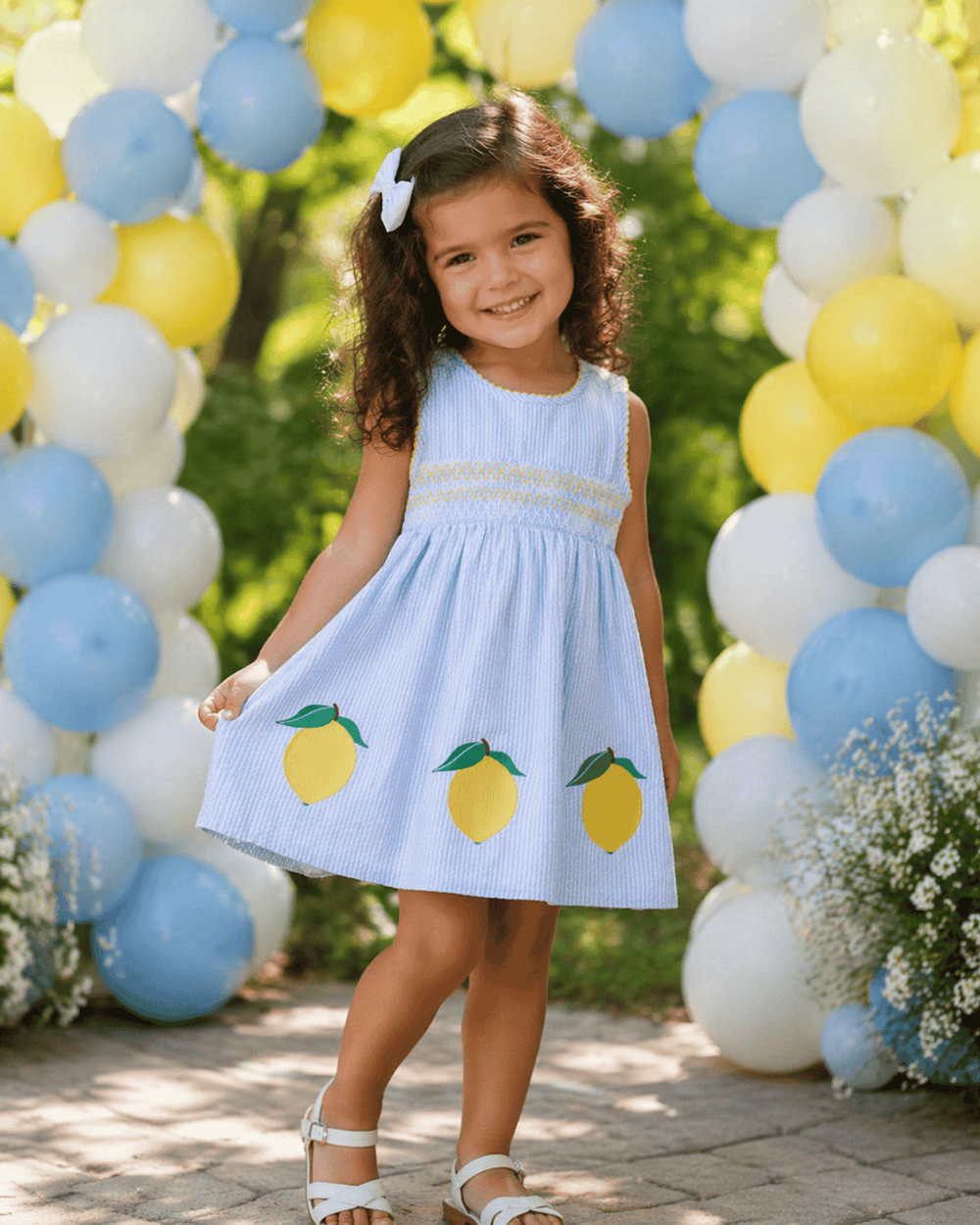 Young girl in a light blue dress with lemon designs standing in front of balloons.