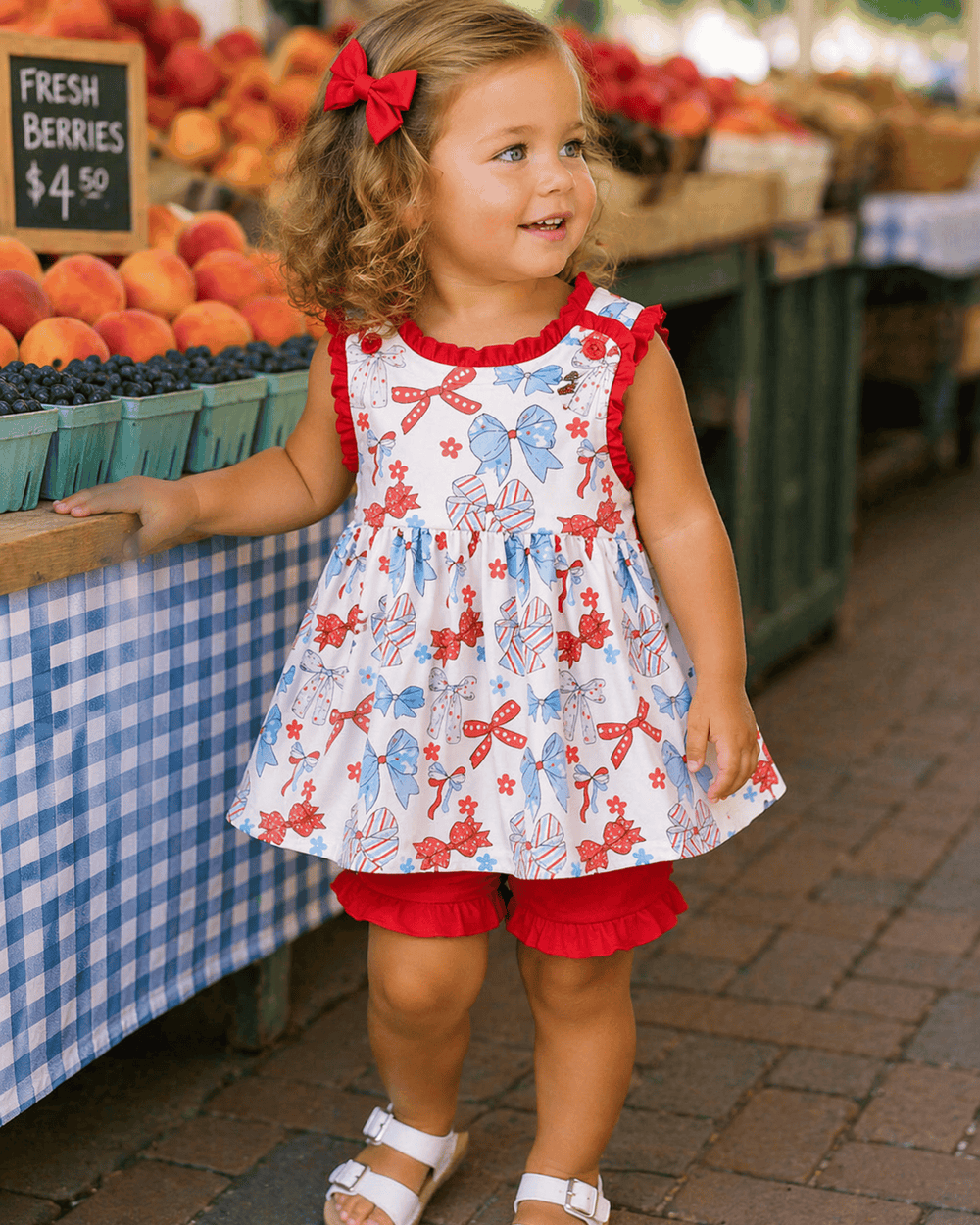 Young girl in a colorful dress standing at an outdoor market with fruits and vegetables in the background.