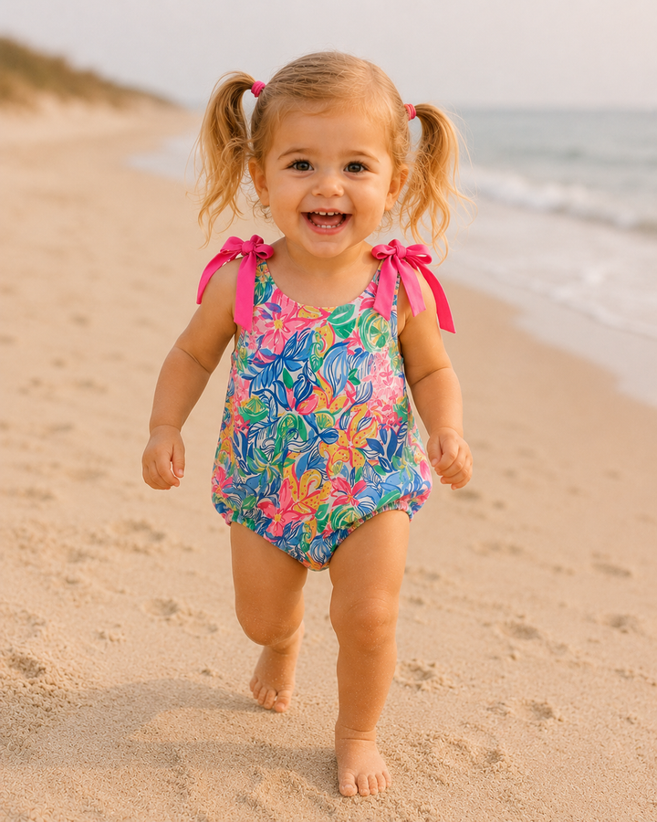 Child in a colorful swimsuit with floral pattern and pink bows running on a sandy beach.