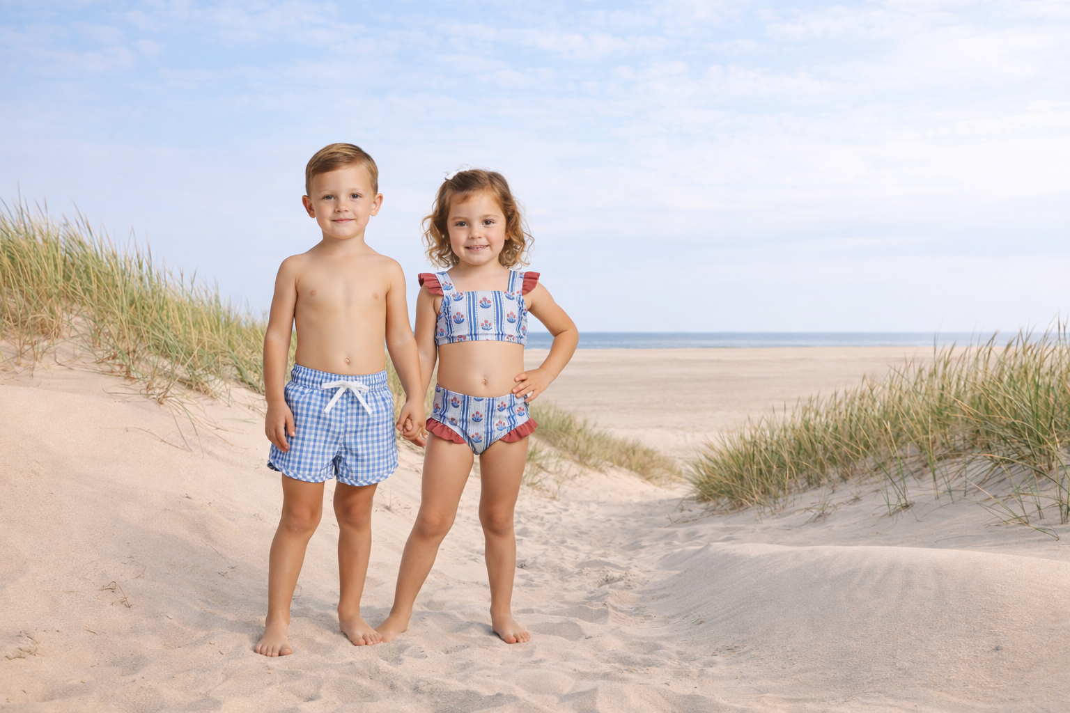 Two children in matching outfits standing on a sandy beach with grassy dunes and blue sky.