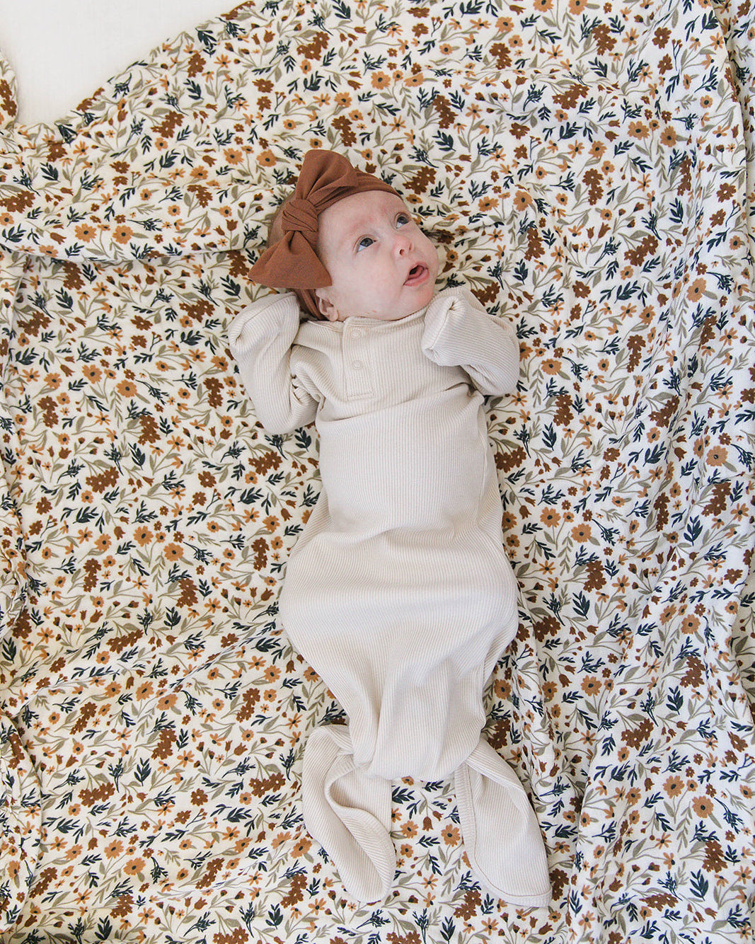 Baby lying on a floral blanket wearing a white outfit and brown hat.