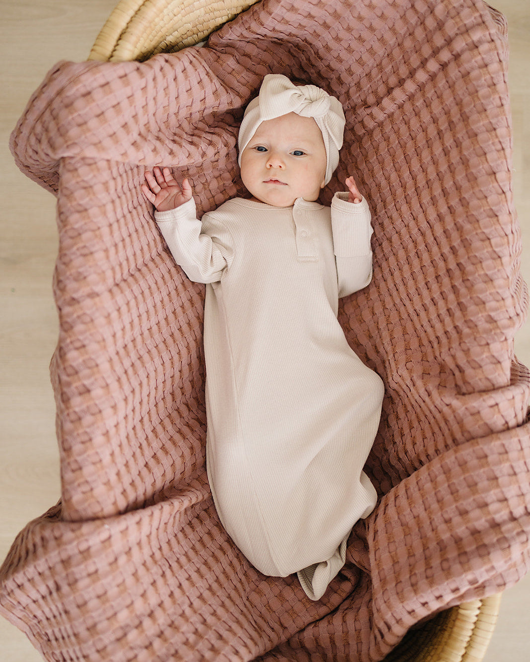 Baby in a white outfit lying on a pink textured blanket in a wicker basket.