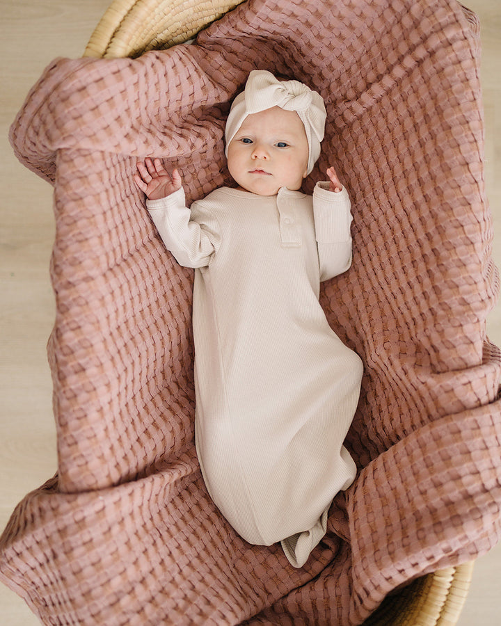 Baby in a white outfit lying on a pink textured blanket in a wicker basket.