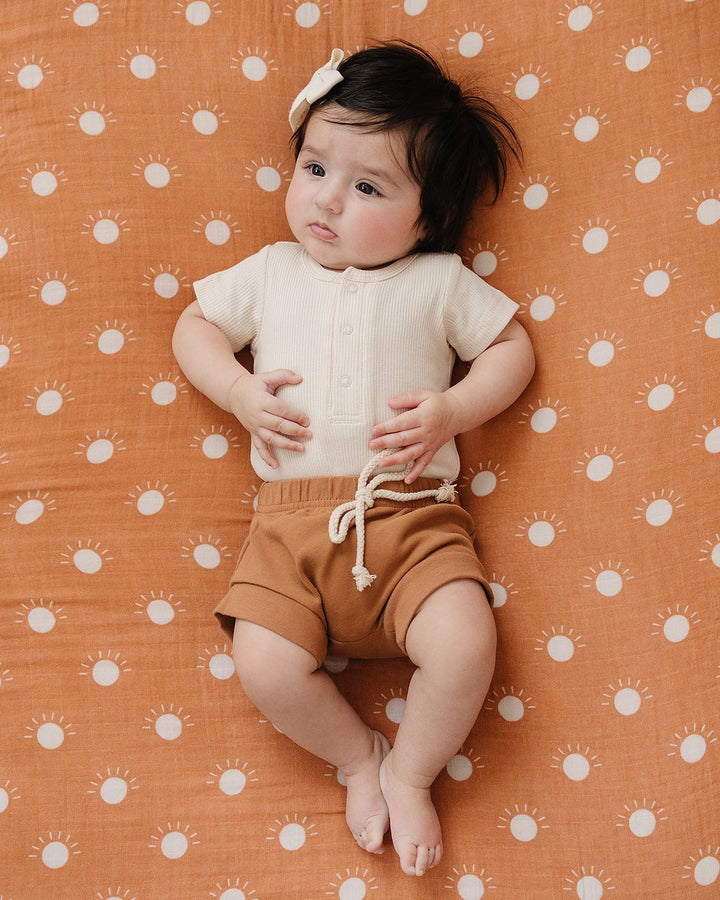 Child laying on a patterned orange surface wearing a beige shirt and brown shorts.