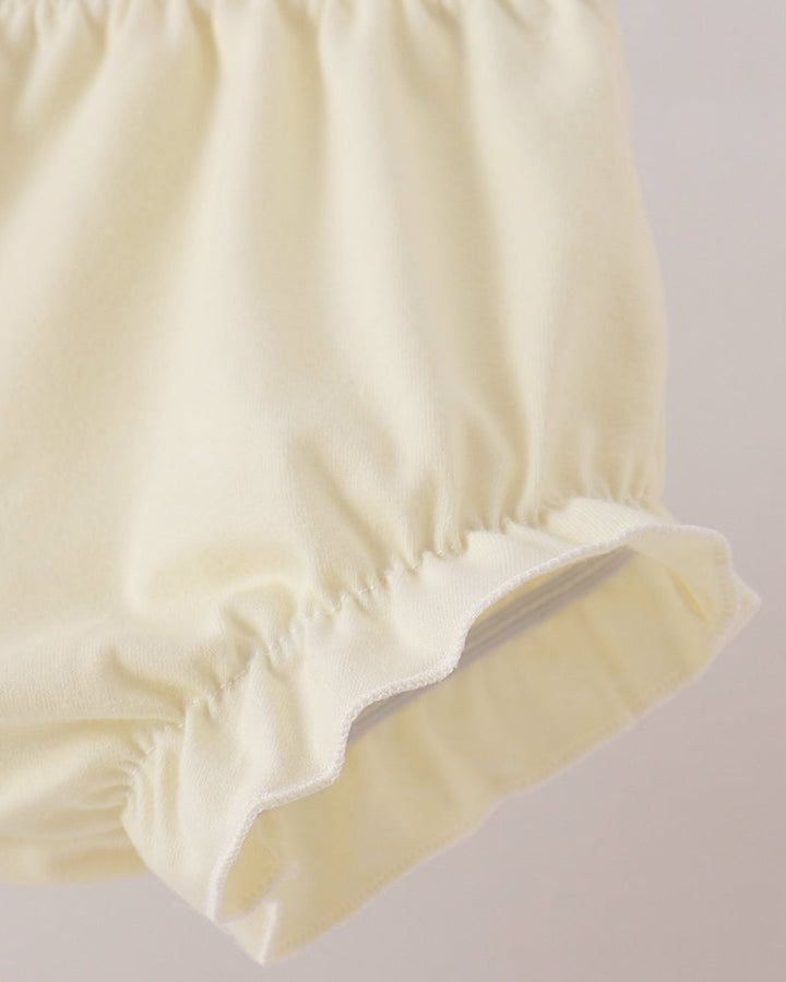 Close-up of a pair of cream-colored ruffled bloomers or diaper cover, showing the elastic waistband and scalloped hem against a light neutral background.