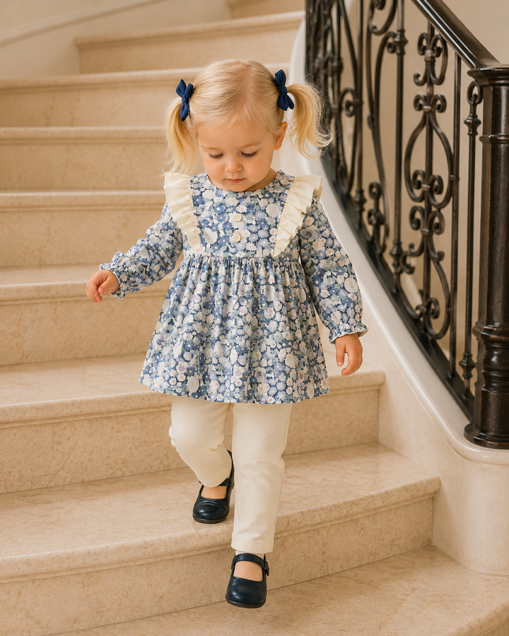 A toddler girl with blonde pigtails wearing a blue and white floral long-sleeve dress, cream pants, and navy shoes walks down a beige stone staircase next to a black wrought-iron railing.
