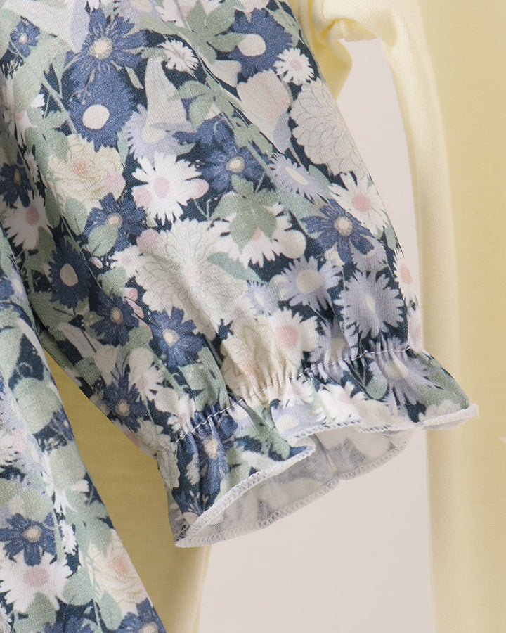 Close-up of a floral-patterned blouse with a ruffled elbow-length sleeve, featuring blue, white, and green flowers on a light background, layered over a pale yellow garment.