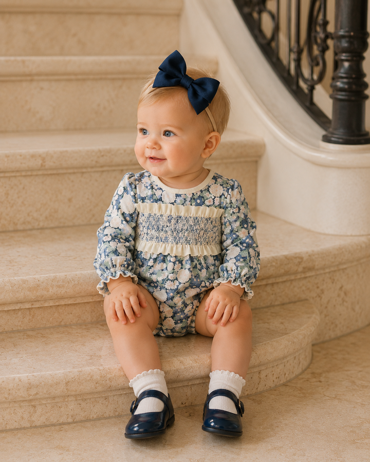 A baby girl sitting on beige stone stairs wearing a blue and cream floral smocked romper, a navy blue bow headband, white socks, and navy shoes.
