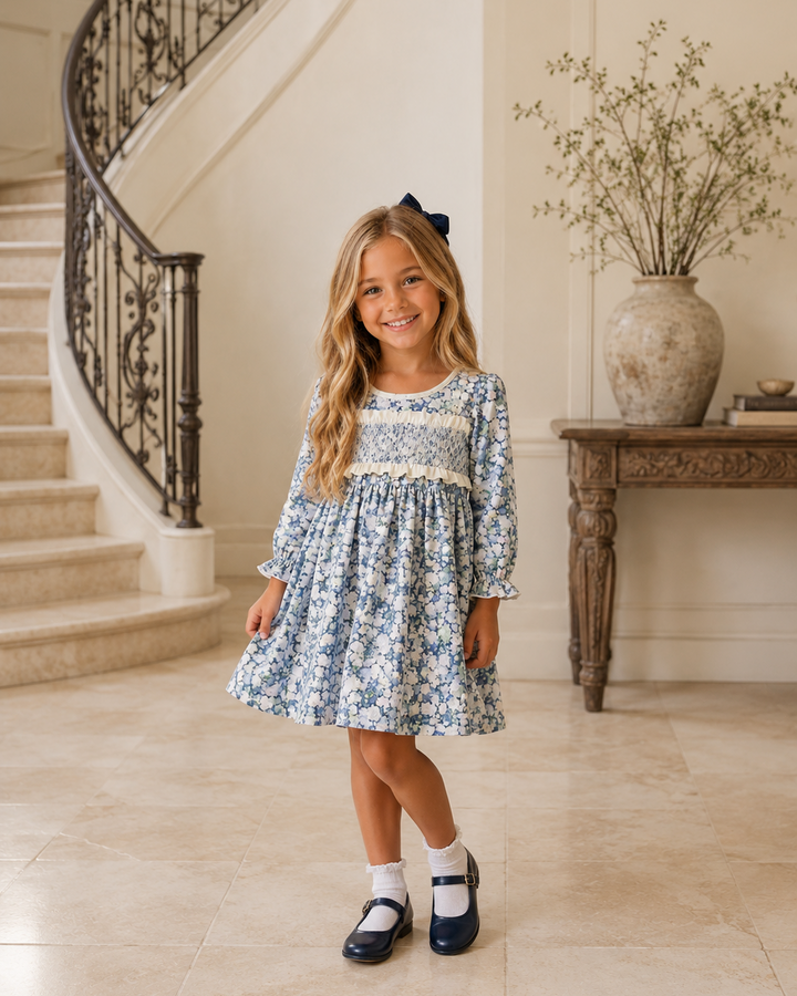 A young girl with long blonde hair stands in a grand staircase hall wearing a blue and white floral print long-sleeve dress with a smocked bodice, white socks, and navy Mary Jane shoes.