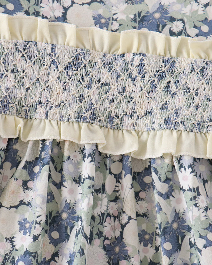 Close-up of a floral dress with a blue, white, and pale green floral print, featuring a cream ruffled band and a smocked cream-and-blue waistband.