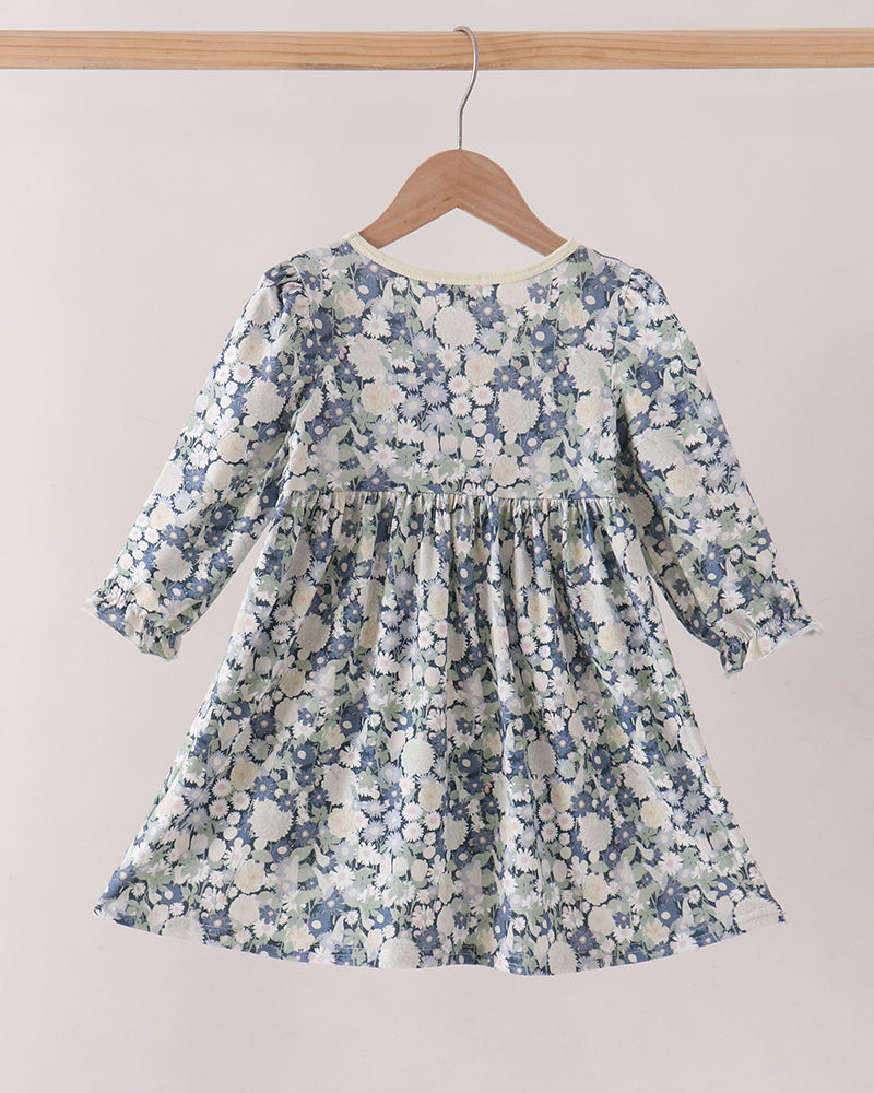 A long-sleeve dress with a blue and white floral pattern, hanging on a wooden hanger from a light wood rod against a plain beige wall.