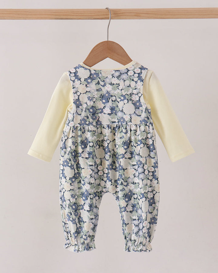 A baby romper with a blue and white floral print over a pale yellow long-sleeve shirt, hanging on a wooden hanger from a light wood rod against a plain beige background.