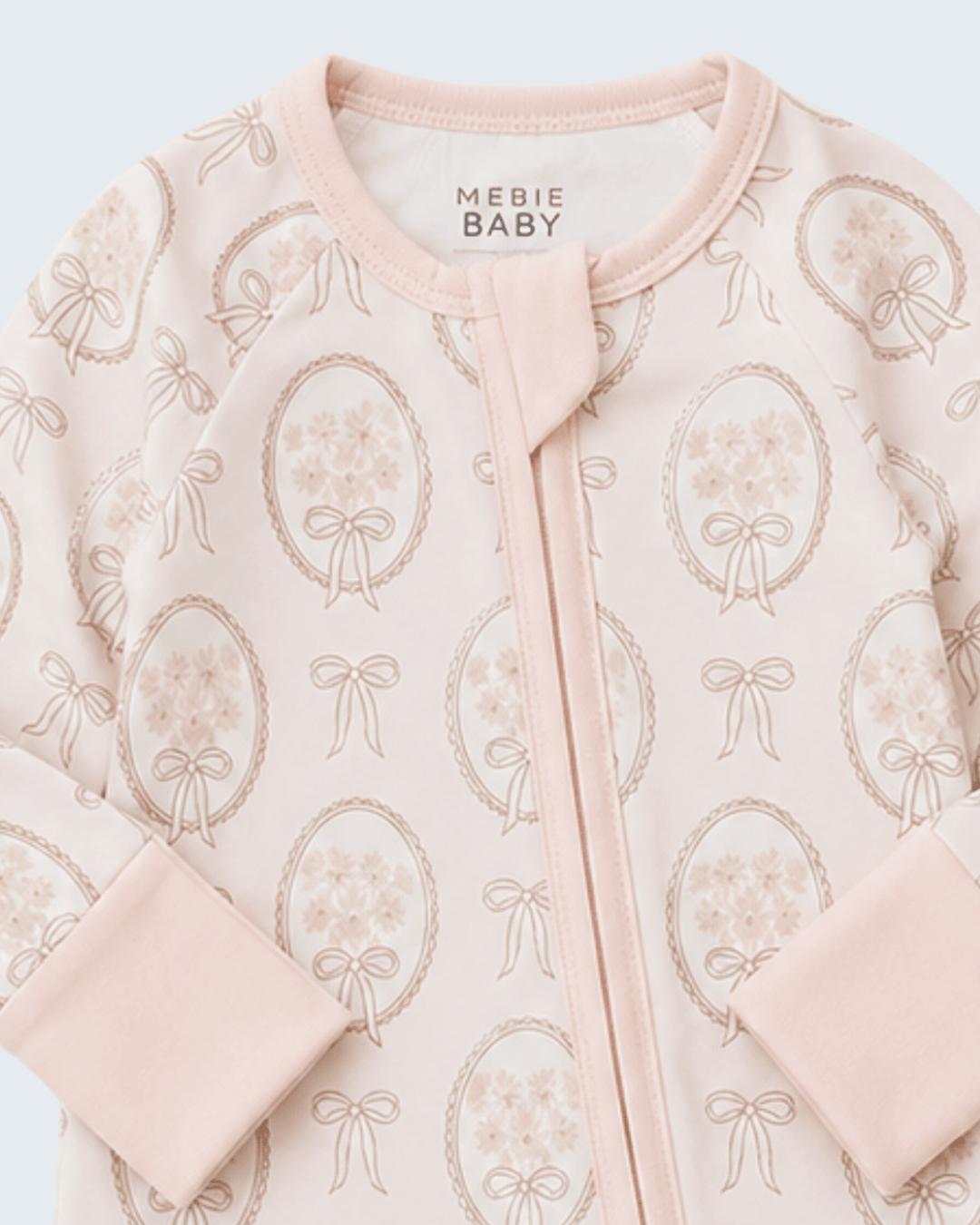 A close-up of a baby sleep sack or long-sleeve top by Mebie Baby, featuring a pale pink base with a repeating pattern of dusty rose bouquets in oval frames and ribbon bows, finished with a matching pink zipper and ribbed collar.