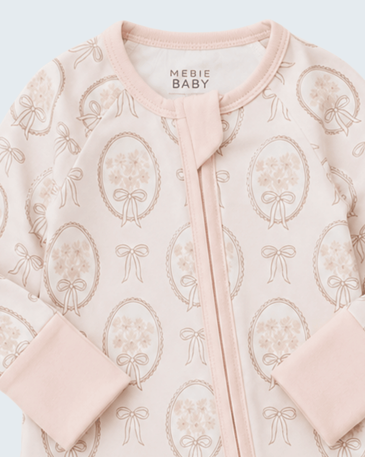 A close-up of a baby sleep sack or long-sleeve top by Mebie Baby, featuring a pale pink base with a repeating pattern of dusty rose bouquets in oval frames and ribbon bows, finished with a matching pink zipper and ribbed collar.