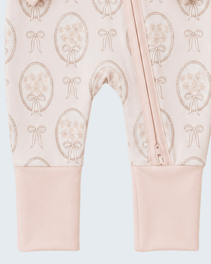 Close-up of a baby footed sleeper or romper in pale blush pink with an allover beige floral oval and bow pattern, plus a front zipper and ribbed cuffs.