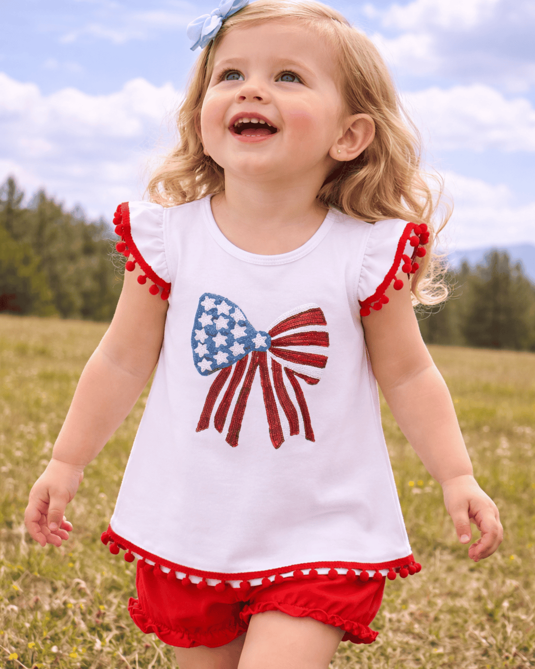 Child wearing a white top with a red, white, and blue flag design and red shorts in a field.