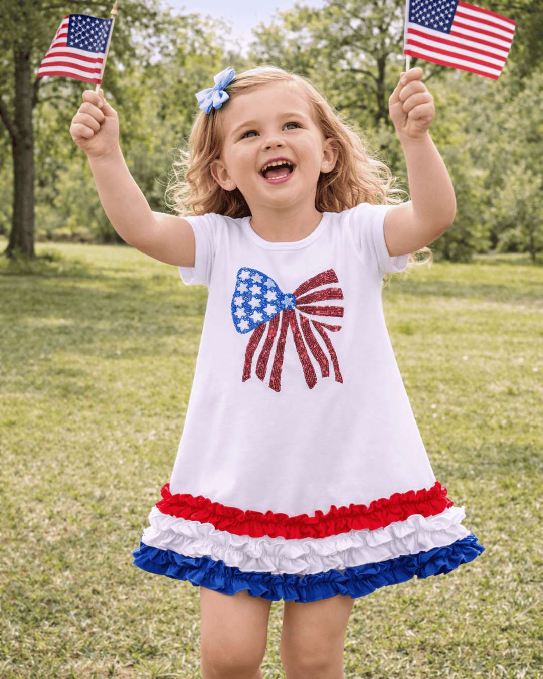 Child wearing a dress with an American flag design, holding two small American flags outdoors.