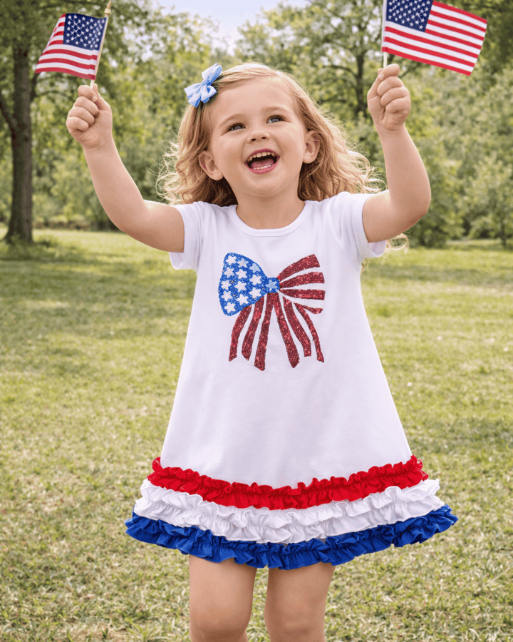 Child wearing a dress with an American flag design, holding two small American flags outdoors.