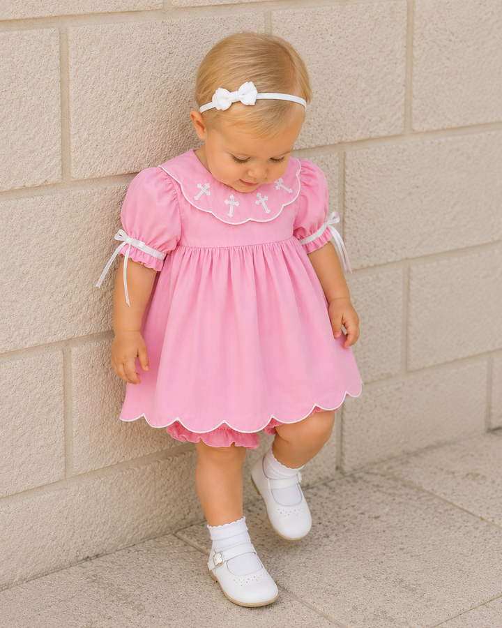 A toddler girl wearing a pink dress with a white bow headband, white socks, and white shoes, standing against a light beige stone wall.