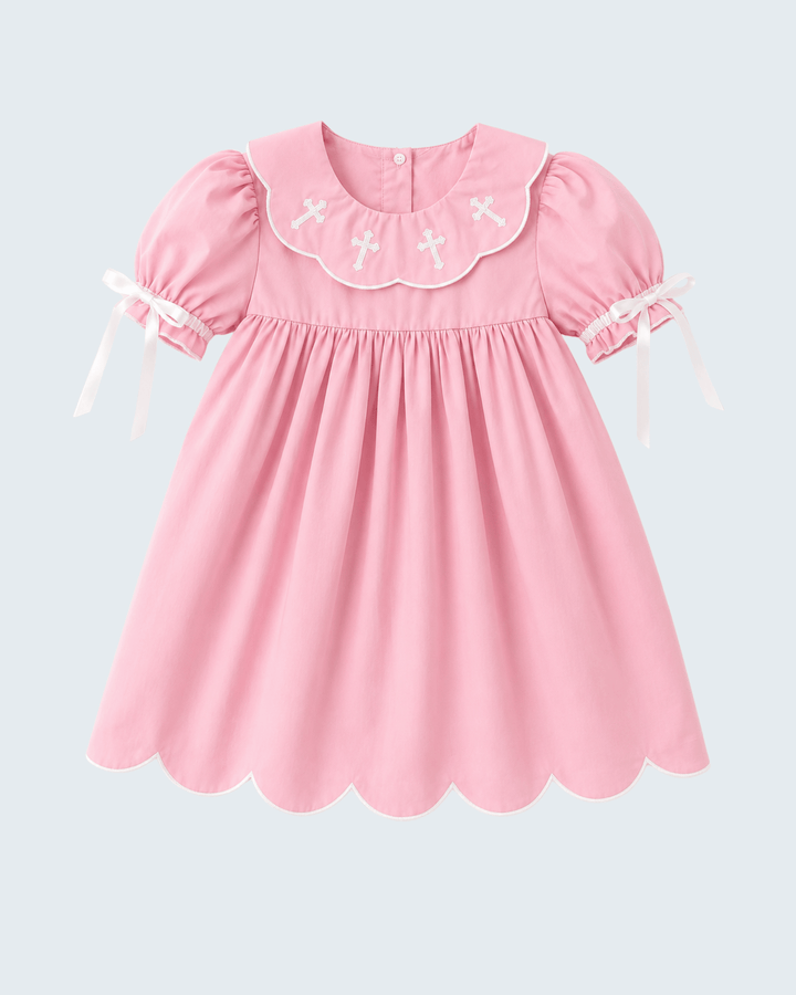 A pink short-sleeve dress with a scalloped Peter Pan collar decorated with white embroidered crosses, featuring puffed sleeves with white ribbon ties, shown against a light gray background.