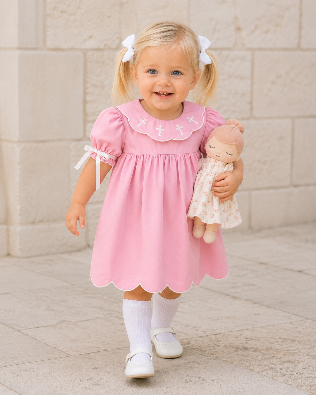 A young blonde girl with pigtails wearing a pink dress with white bows and white socks, holding a doll while standing outdoors against a light stone wall.